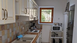 Une cuisine compacte avec des meubles blancs, un plan de travail en bois, une fenêtre à barreaux, un four à micro-ondes et une poubelle.