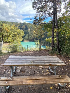 Ein Holztisch mit Metallbeinen, vor einer bewaldeten Hügelslandschaft und einem türkisblauen Gewässer.