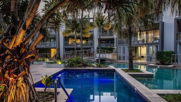A modern hotel with a swimming pool, palm trees, and balconies.