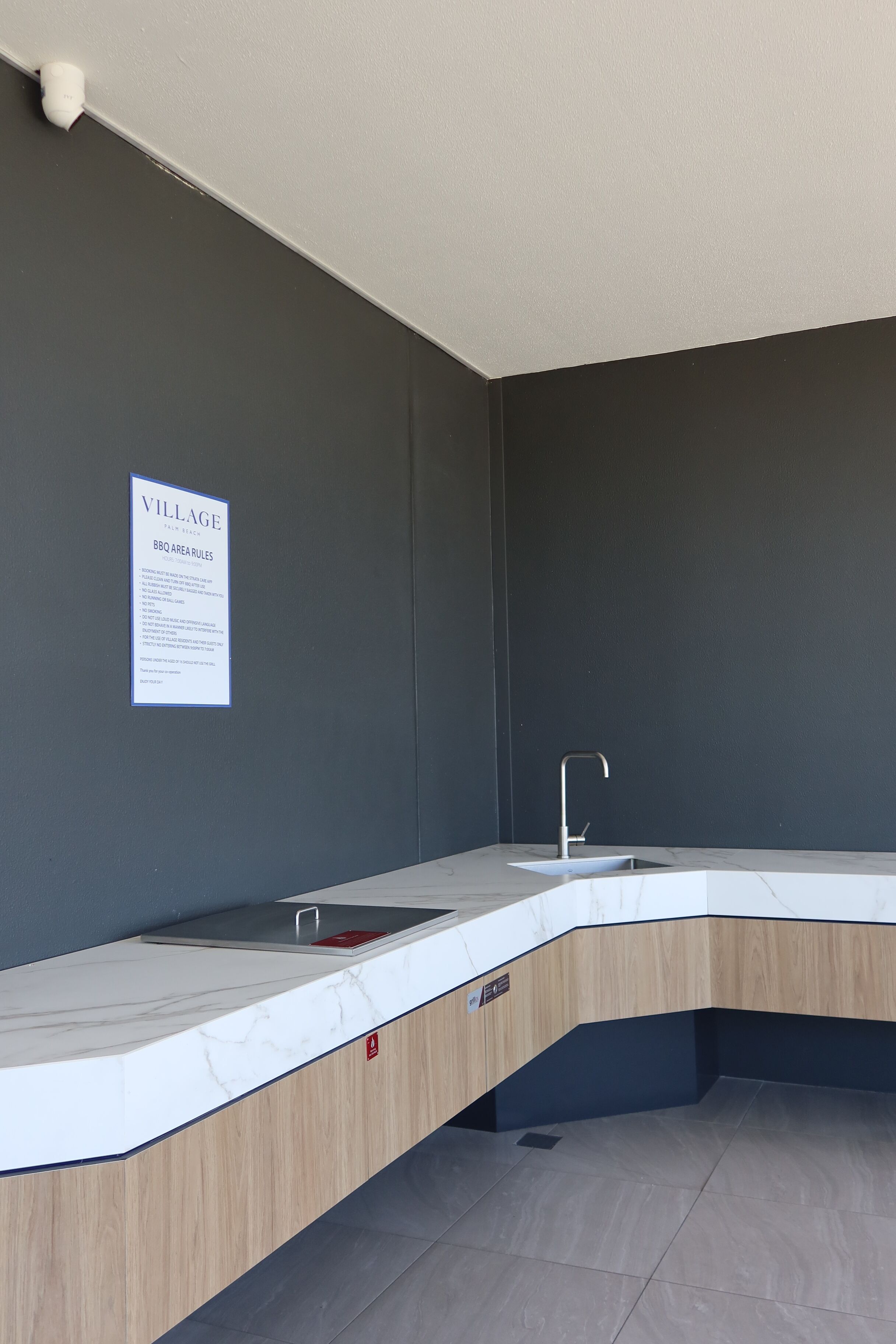 A modern bathroom with a marble countertop, a sink, and a wooden cabinet. There is a sign on the wall titled "Village BBQ Area Rules".