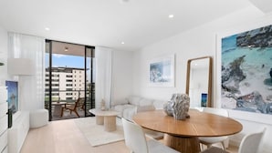 A modern living room with a round wooden table, white chairs, a sofa, and a large window with a view of buildings and a beach. - Luxury Palm Beach 1 BDR in The Village (Palm Beach)