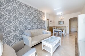 A living room with a patterned wallpaper, a beige sofa, a glass coffee table, and a dining area with chairs and a table. - Calanova Grand Golf stunning apartment (Mijas Costa)
