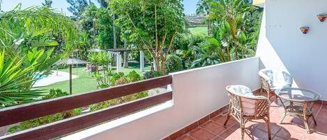 Un balcon doté d'une balustrade en bois, de meubles en osier et offrant une vue sur un jardin luxuriant, avec une variété de plantes et une piscine.