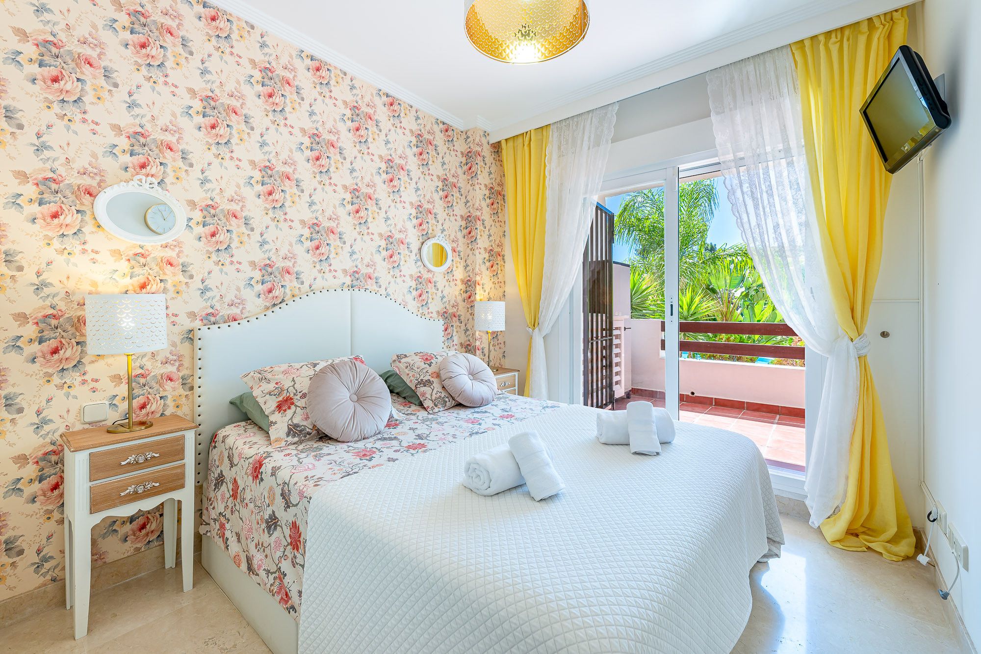 A bedroom with floral wallpaper, a bed with white bedding, a bedside table with a lamp, and a TV mounted on the wall.
