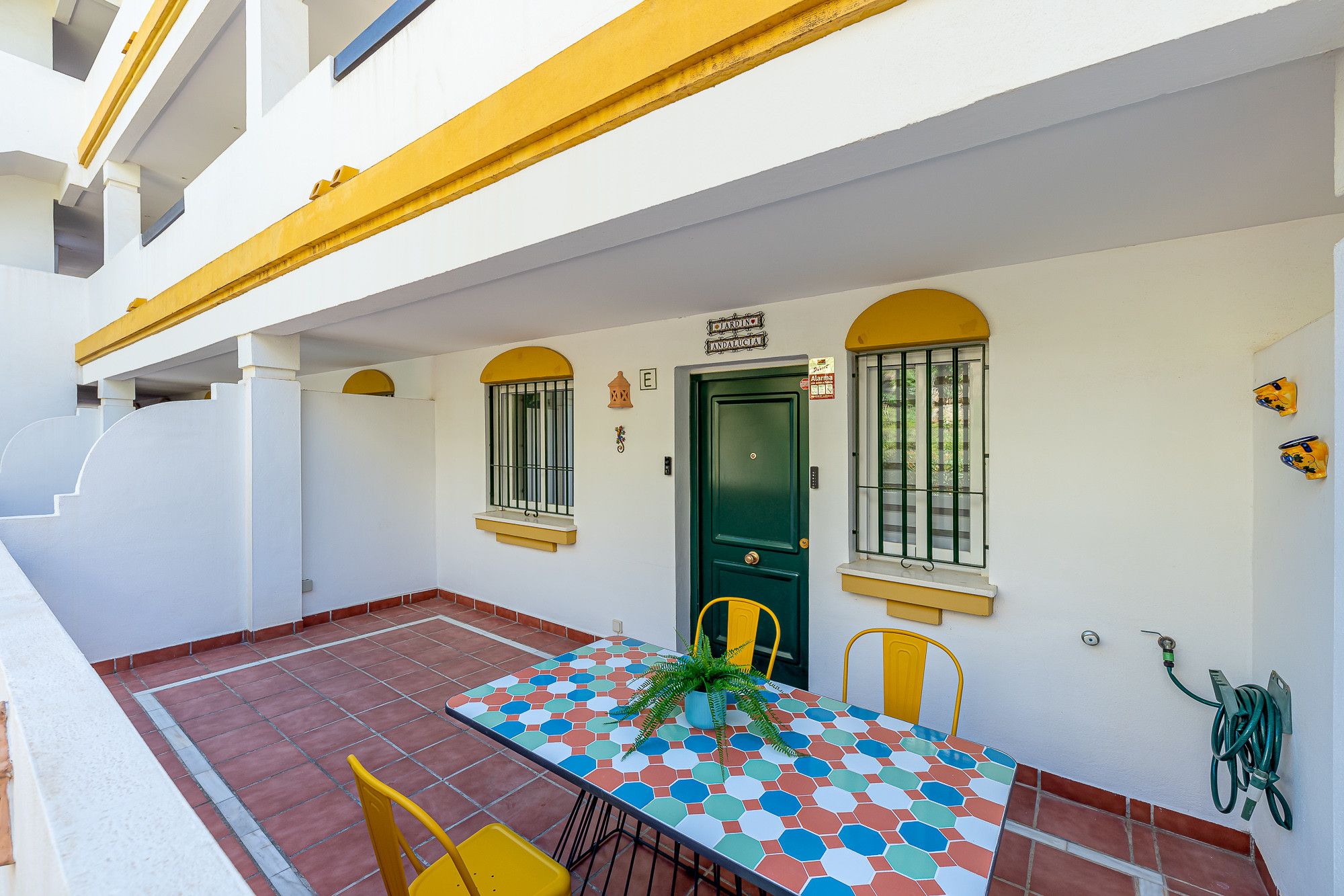 A building with a green door, yellow-framed windows, and a tiled patio area with a table and chairs.