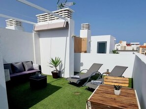 A rooftop terrace with a green artificial turf, a sofa, two lounge chairs, a small wooden table, and a potted plant.
