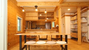 Un interior de madera con una mesa larga y sillas, una zona de cocina con armarios y un refrigerador, y un estante con cestas.