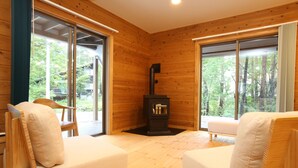 Una cabaña acogedora con paredes de madera, chimenea, dos sillones mullidos y ventanas amplias con vistas al bosque.