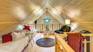 Um quarto de sótão aconchegante com teto de madeira, uma pequena janela, uma cama com almofadas vermelhas e brancas, um banco de madeira e um tapete redondo.
