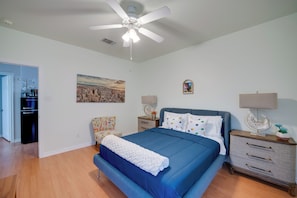 Une chambre à coucher comprenant un lit, des tables de chevet, un ventilateur de plafond, un tableau représentant un paysage urbain et une porte donnant sur une autre pièce.