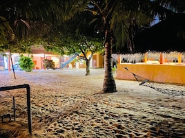 Uma área de praia arenosa com uma rede, palmeiras e um prédio com escadaria.