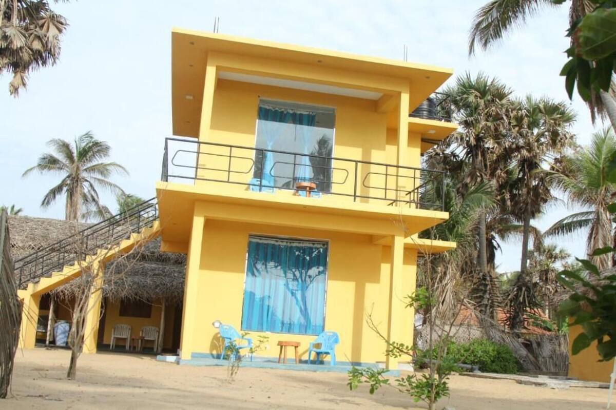 A two-story yellow building with a balcony, a staircase, and a small outdoor seating area with chairs.