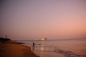 Cena de praia ao anoitecer com uma pessoa pescando, um píer ao fundo e estruturas industriais na água.