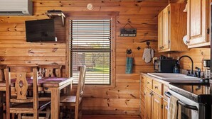 Interior de una cabaña con paredes de madera, una zona de comedor con sillas y una mesa, una cocina con armarios y electrodomésticos, y una ventana con persianas.