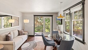 Un salón moderno con una mesa de comedor de cristal, sillas grises, un sofá beige con cojines, una alfombra redonda y ventanas amplias con vistas al verdor.