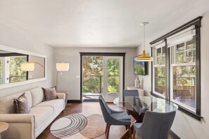 A modern living room with a glass dining table, grey chairs, a beige sofa with cushions, a round rug, and large windows with a view of greenery.