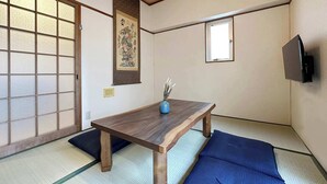 Ein traditioneller japanischer Raum mit Tatami-Boden, einem niedrigen Holztisch, einem blauen Kissen, einem an der Wand montierten Fernseher und einer Shoji-Schiebetür.