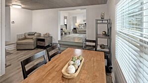 Un espace repas avec une table en bois, des chaises et un centre de table composé d’objets décoratifs. À l’arrière, on aperçoit un coin salon avec des canapés et une table basse.