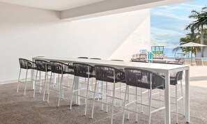 A modern outdoor dining area with a white table and black chairs, a pool, and palm trees in the background.