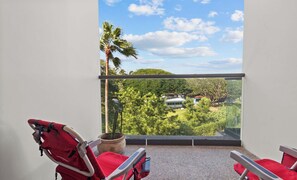 A balcony with two red chairs, a potted plant, and a view of a green landscape with trees and a building.