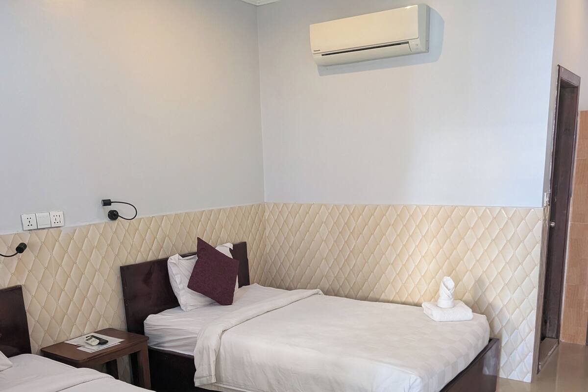 A hotel room with two beds, a wall-mounted air conditioner, and a bedside table with a phone and a notepad.
