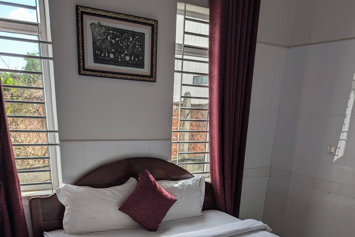 A neatly made bed with white linens and a single red pillow, a framed artwork on the wall, and a window with curtains.