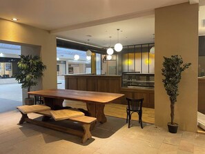 Enjoy meals in this dining area with a wooden table and benches, surrounded by potted plants and a kitchen area.