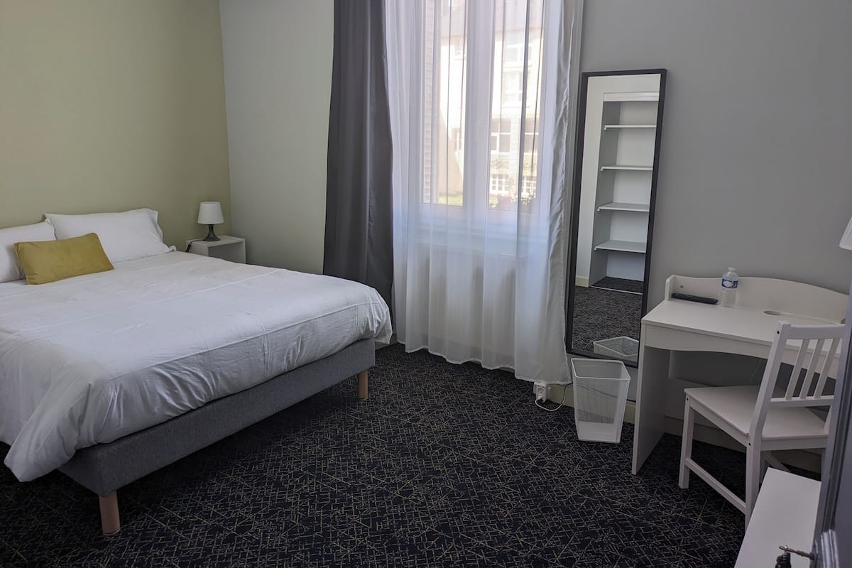 A hotel room with a bed, a desk, a chair, a mirror, a nightstand, and a window with curtains.