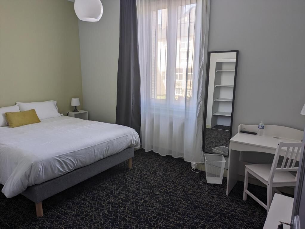Habitación de hotel con cama, escritorio, silla, espejo y ventana con cortinas.