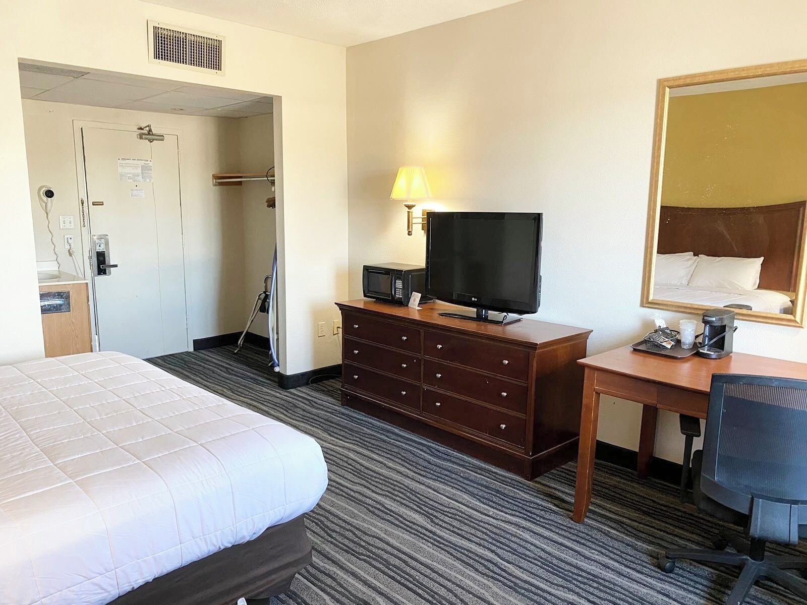 A hotel room with a bed, a dresser, a desk with a computer, a TV, a lamp, and a mirror.