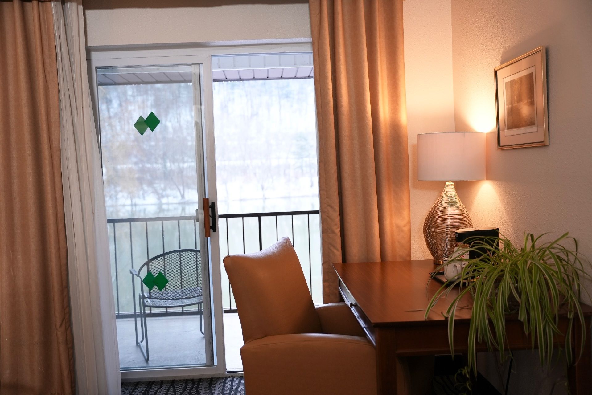 A hotel room with a sliding glass door, a chair, a table with a lamp, and a plant.