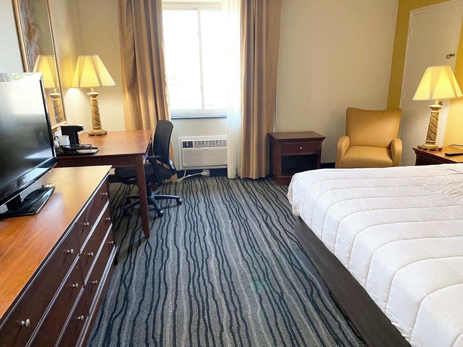 A hotel room with a bed, a desk with a TV, a chair, a lamp, and an air conditioning unit.