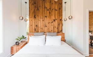 A bedroom with a wooden headboard, a bed with white bedding, two bedside tables with plants, and pendant lights.
