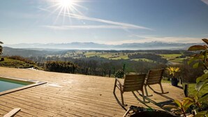 Una terraza de madera con piscina, dos mecedoras y vista a montañas distantes.