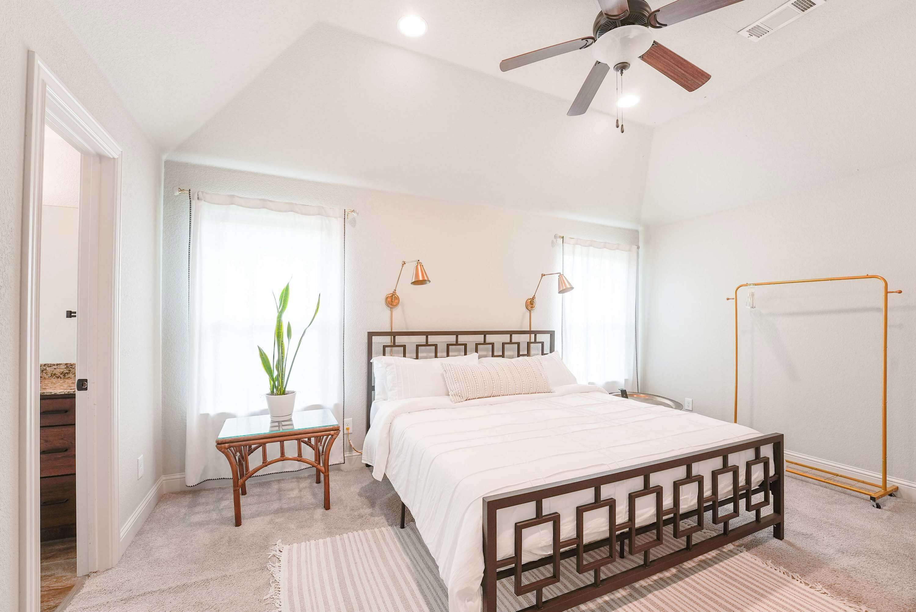 Un dormitorio con cama, ventilador de techo, ventana con cortinas, una mesita con una planta y una lámpara de pie.