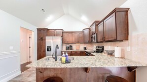 A kitchen with granite countertops, wooden cabinets, a stainless steel refrigerator, a microwave, and a coffee maker. - Modern 3BR Retreat • EV Charger • Fenced Yard by XNA (Cave Springs)