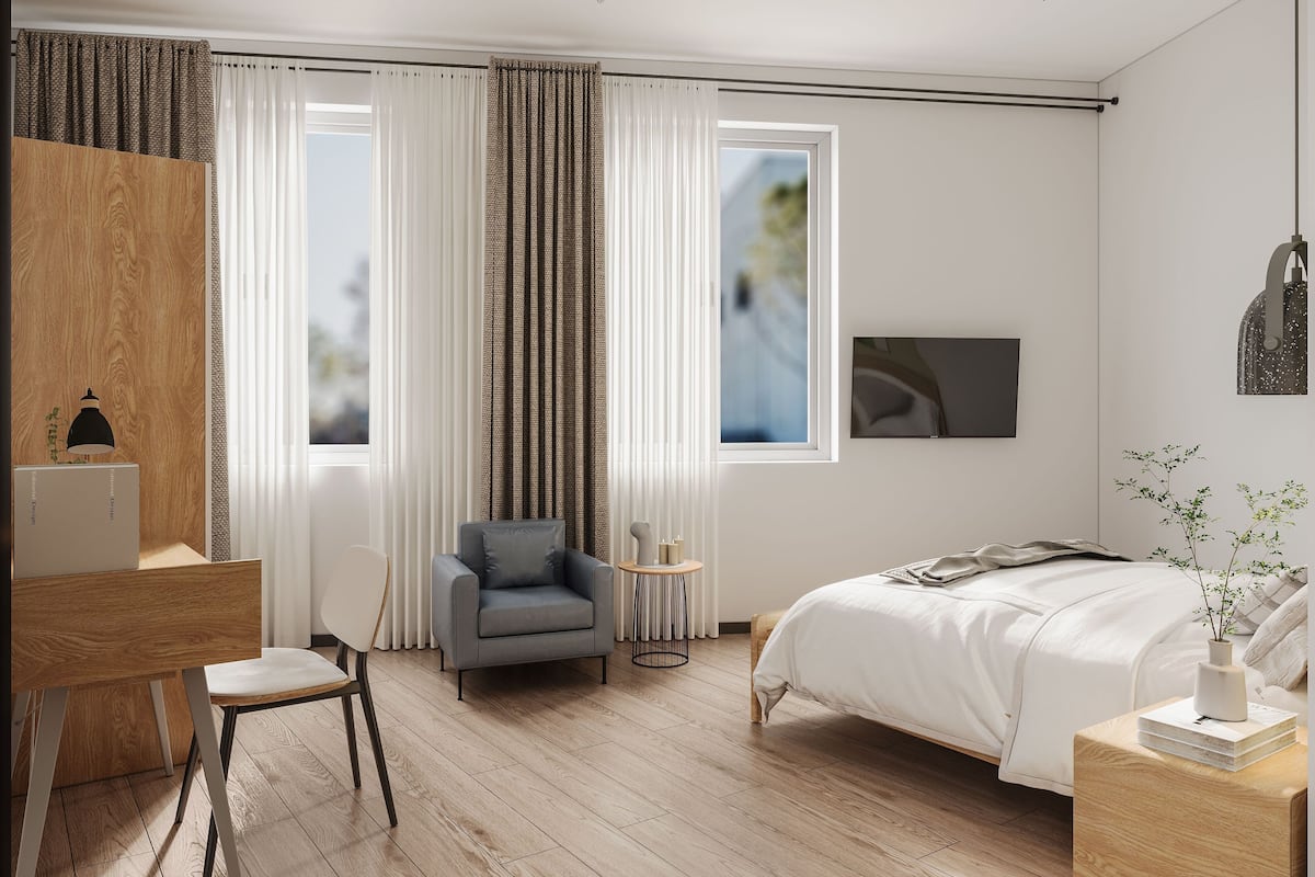 A modern bedroom with a wooden floor, a desk with a lamp, a grey armchair, a bedside table with a vase, and a bed with a white duvet.