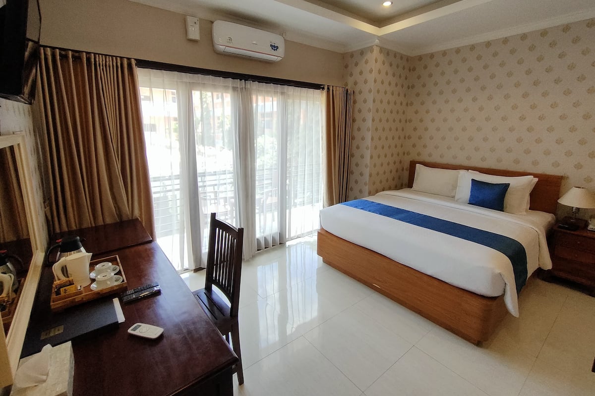 A hotel room with a bed, a desk with a chair, a window with curtains, and an air conditioning unit on the wall.