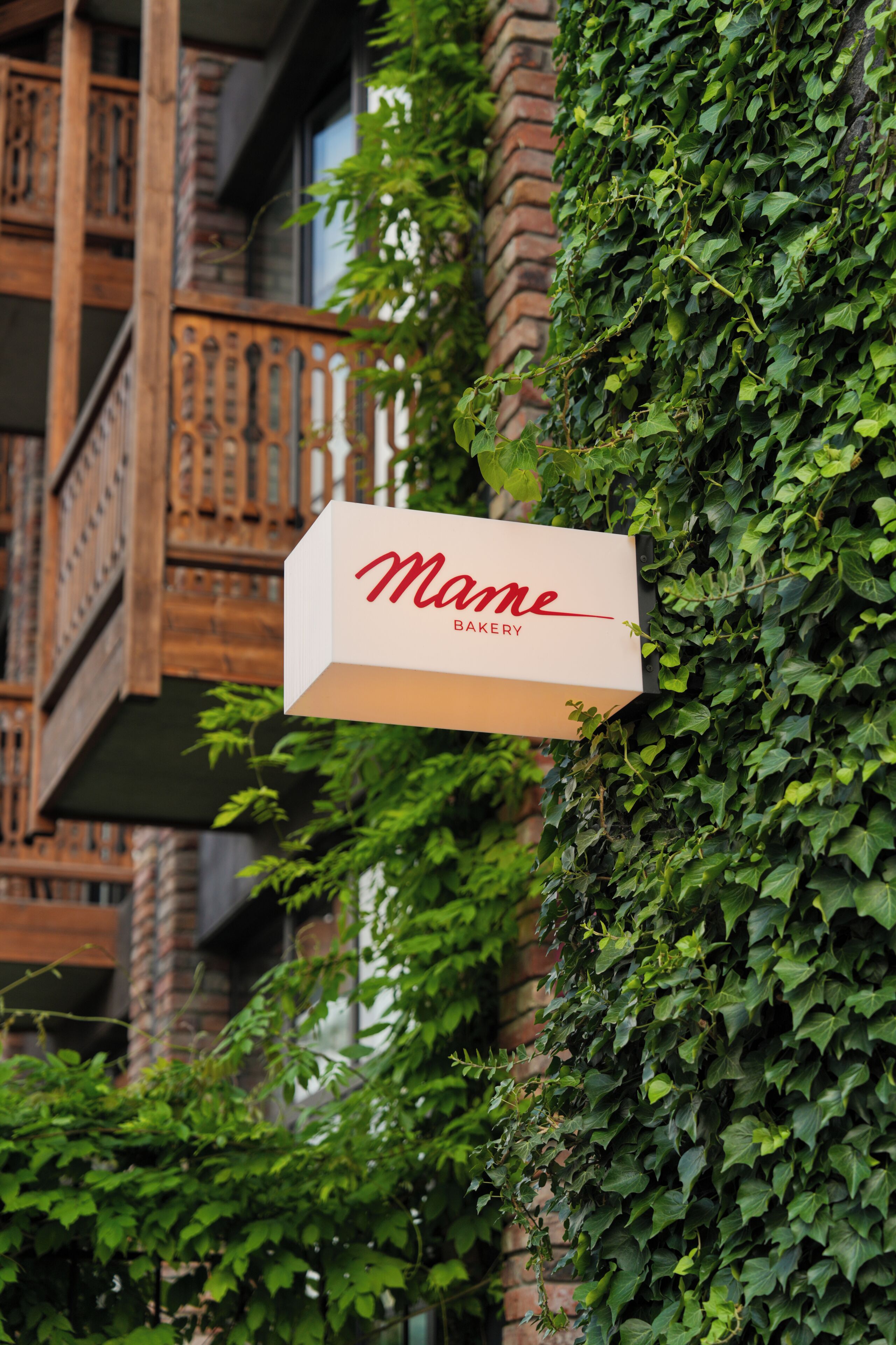 En skylt med texten ”Mame Bakery” sitter fast på en byggnad med en fasad av tegel och en balkong med räcken av trä.