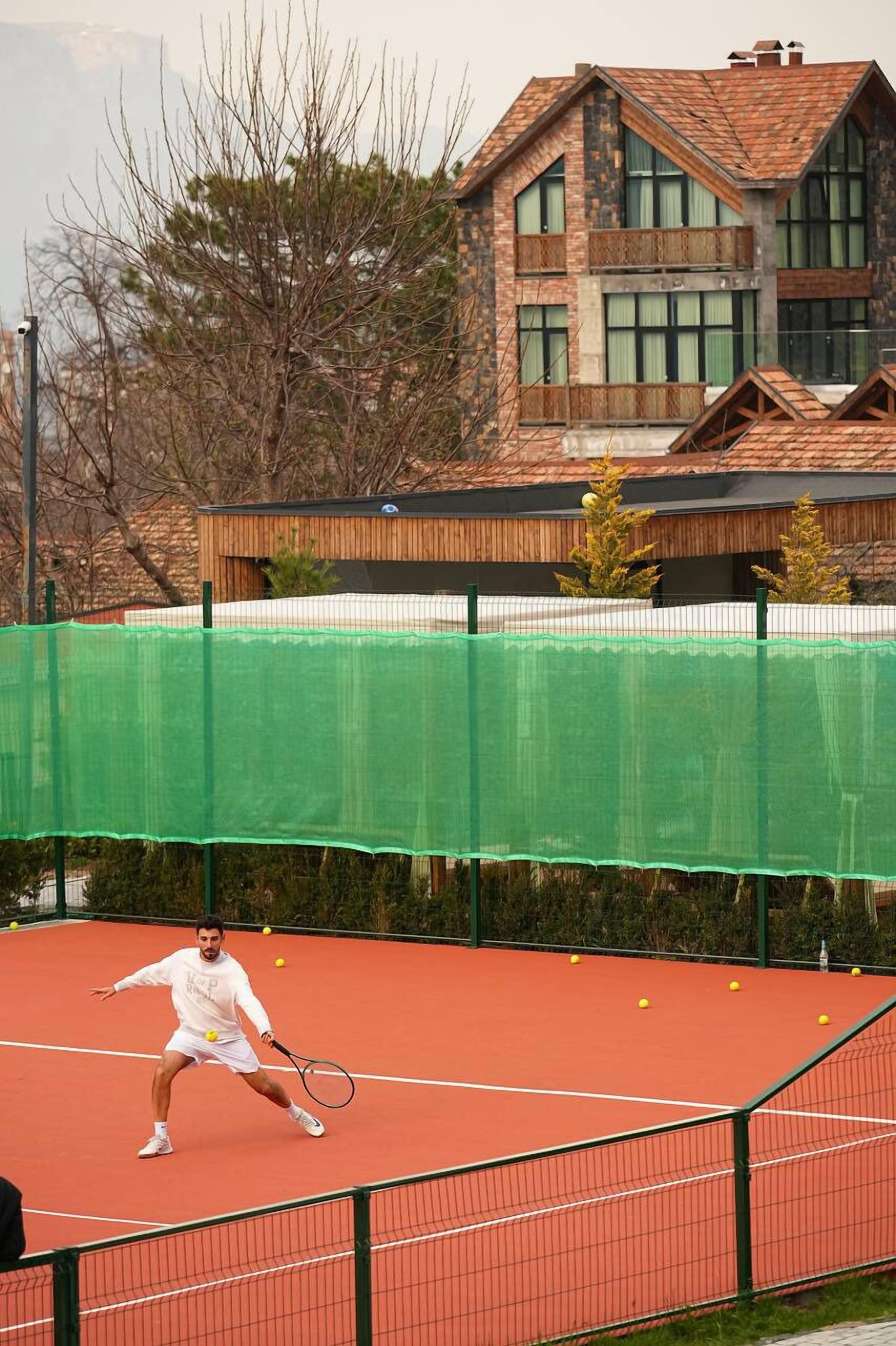 A tennis court with a player in action, surrounded by a green net and a building with a brick facade in the background.