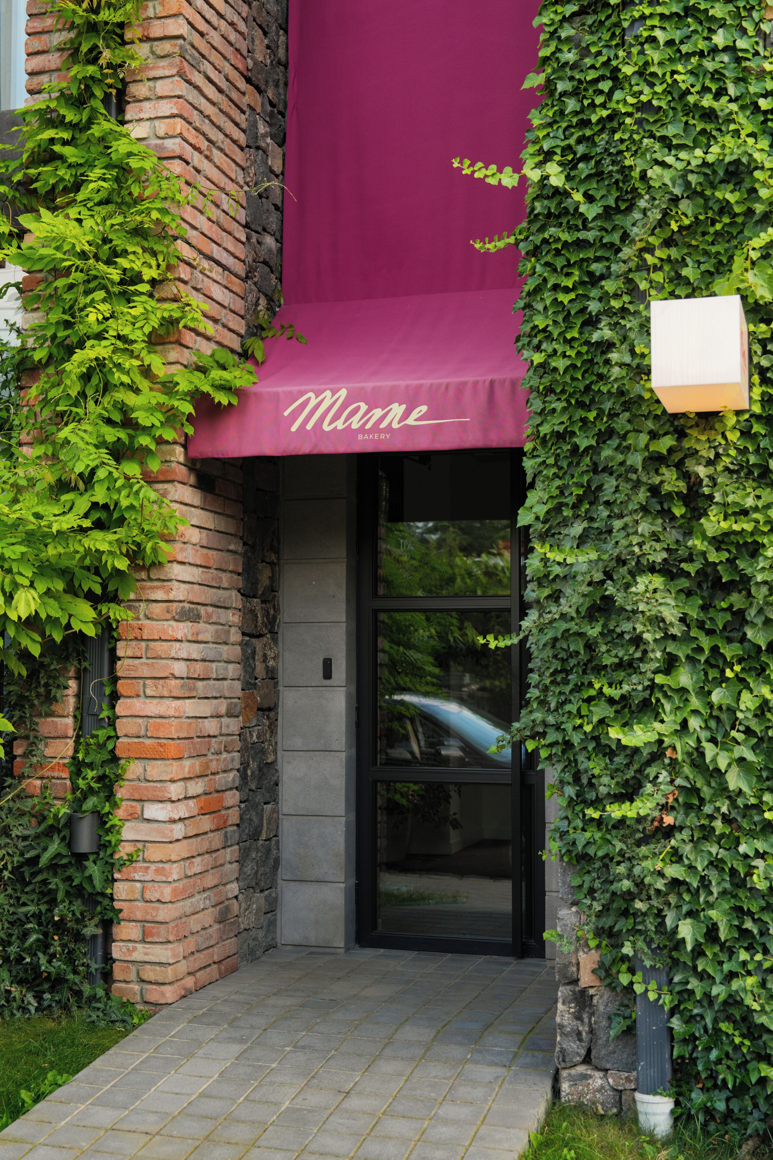 A building entrance with a brick facade, a purple awning with the word 'Mane' and 'Bakery' written on it, and a glass door.