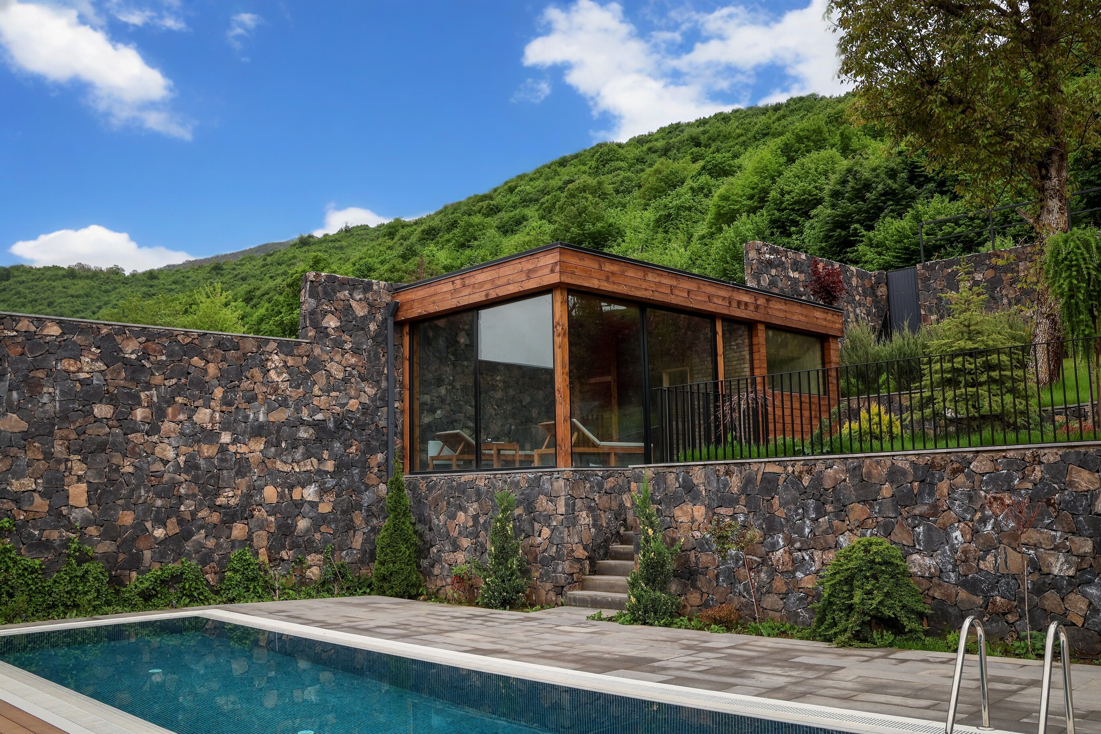 A modern outdoor pool area with a stone wall and wooden deck, surrounded by lush greenery and a mountain backdrop.