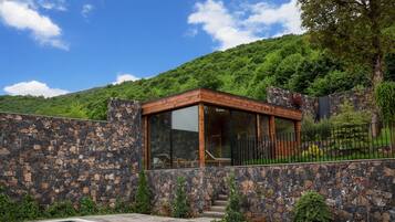 A modern outdoor pool area with a stone wall and wooden deck, surrounded by lush greenery and a mountain backdrop.