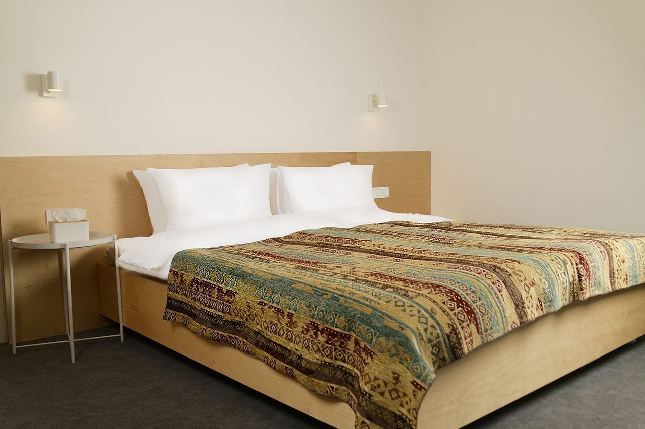 Habitación de hotel con cabecera de madera, cama con ropa blanca, manta decorativa y una mesita de noche pequeña con un dispensador de pañuelos, además de dos luces fijadas a la pared.