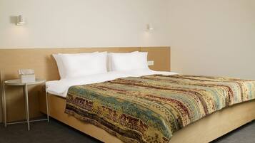 Habitación de hotel con cabecera de madera, cama con ropa blanca, manta decorativa y una mesita de noche pequeña con un dispensador de pañuelos, además de dos luces fijadas a la pared.