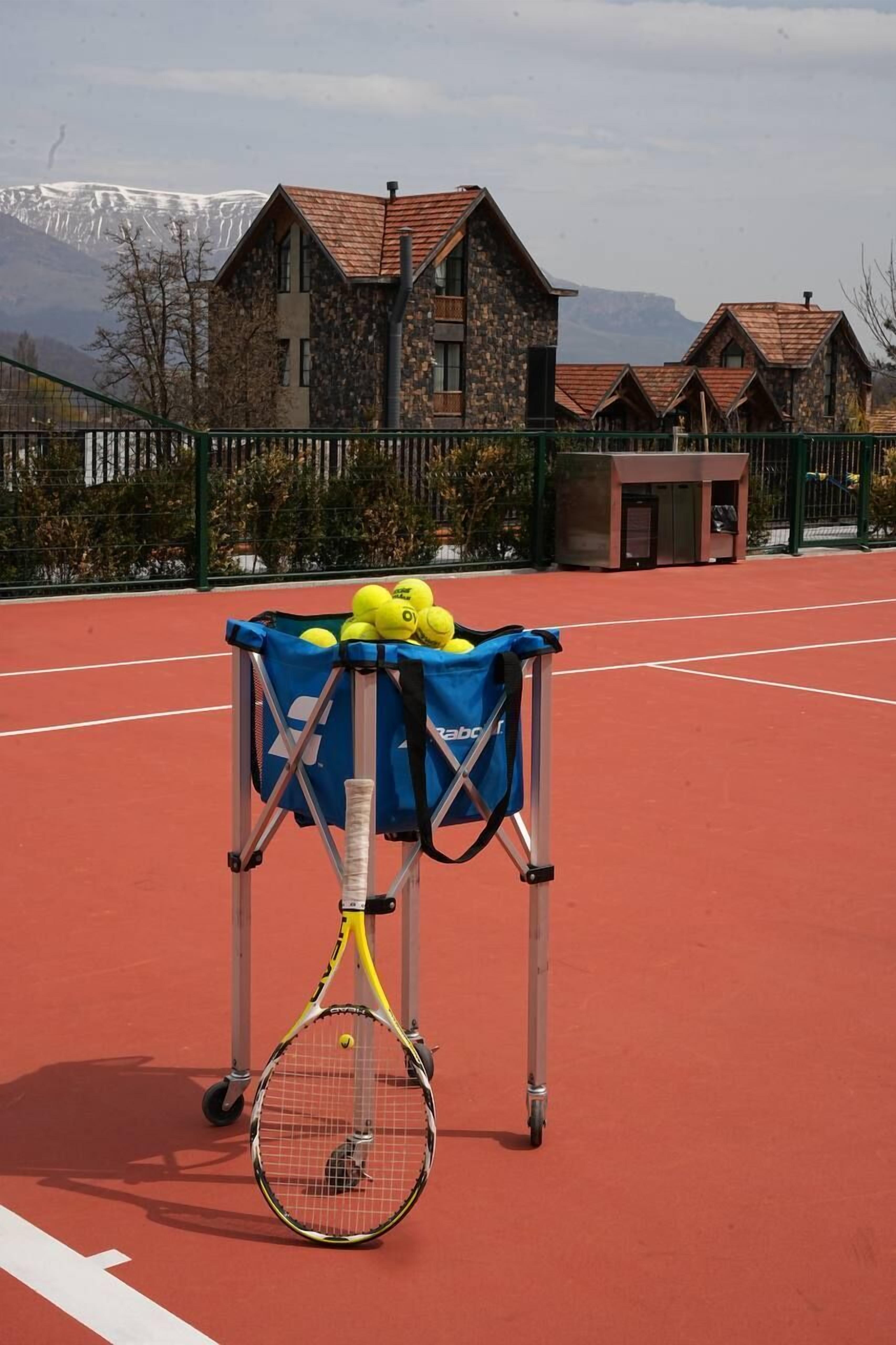 A tennis court with a bag of tennis balls and a racket, a building with a stone facade, and a mountainous backdrop.