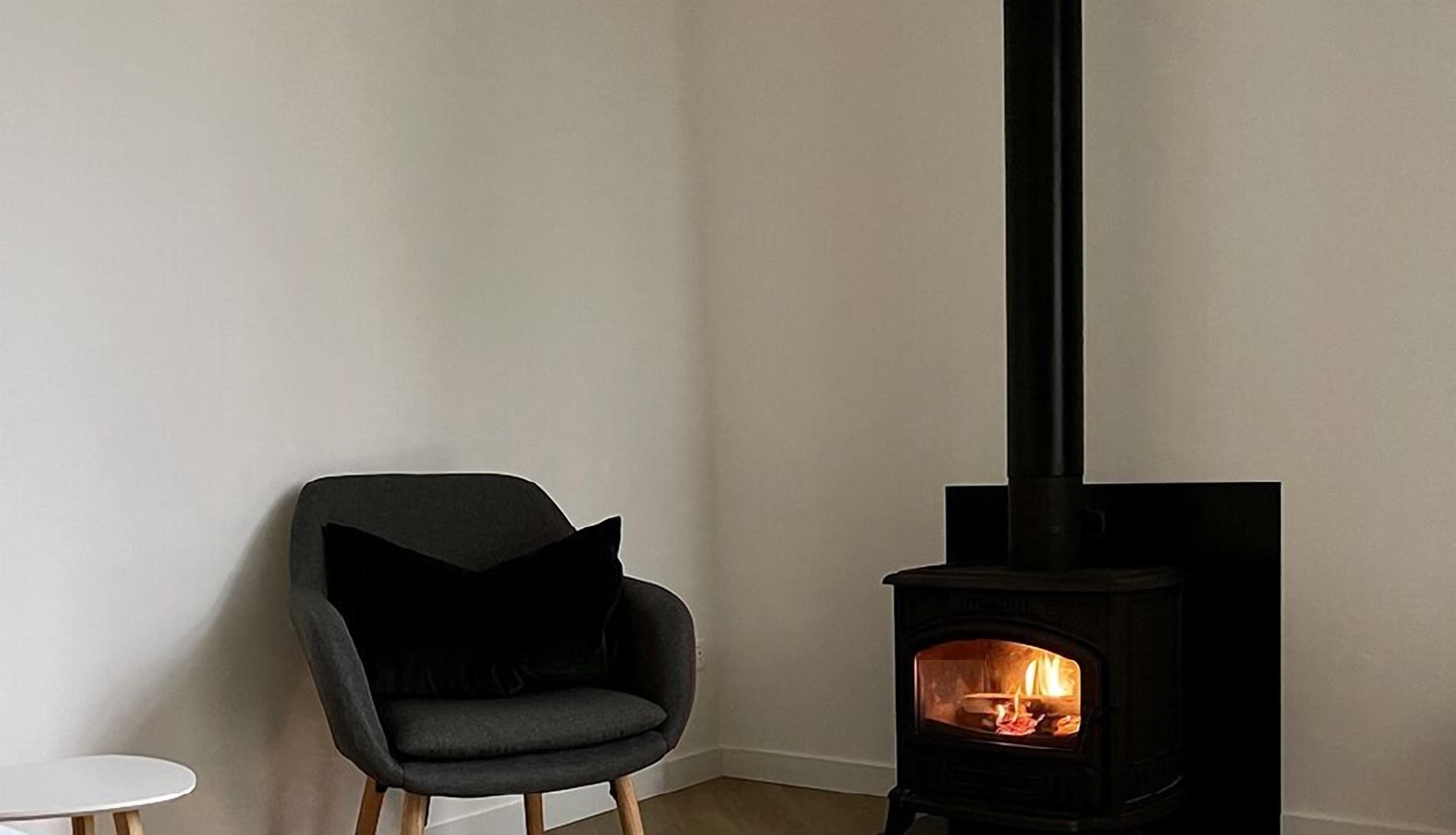 A modern living room with a black wood-burning stove, a grey armchair with a black cushion, and a small white table.