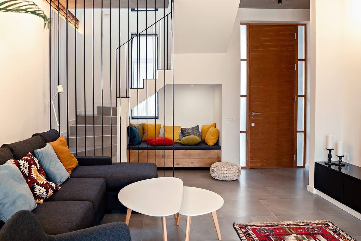 Ein modernes Wohnzimmer mit einer Couch, zwei weißen Tischen, einer Holzbank mit bunten Kissen, einer Treppe mit Metallgeländer und einer Holztür.