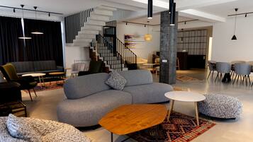 Un intérieur moderne avec un canapé gris, une table basse ronde en bois et un pouf. Un escalier avec des rampes noires mène à un niveau supérieur.