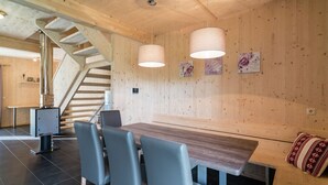 A wooden interior with a staircase, a dining table with chairs, pendant lights, and framed pictures on the wall.